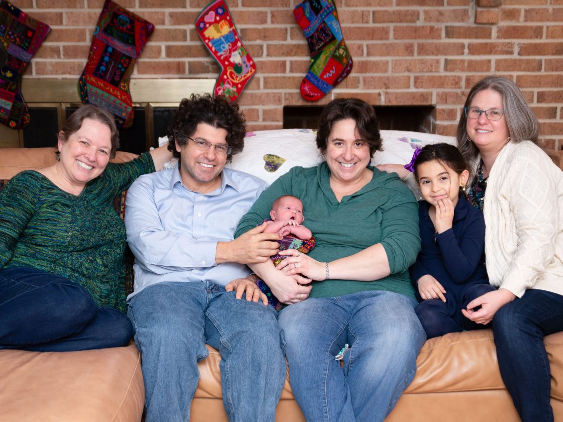 Chris, Melissa, and the Ferraros sit on a sofa in front of Christmas stockings