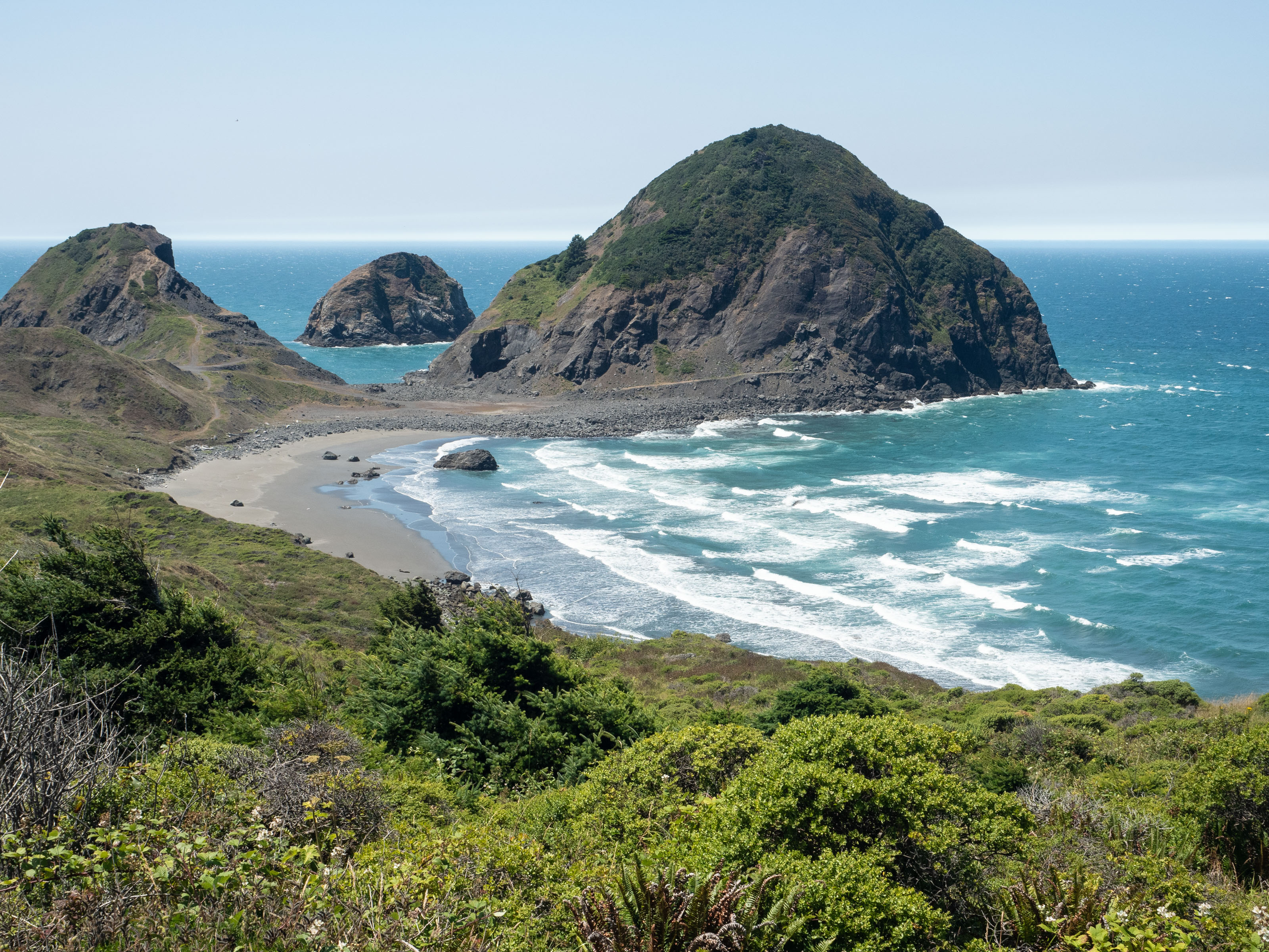 Curve of shoreline with greenery, waves, and triangular sea stacks