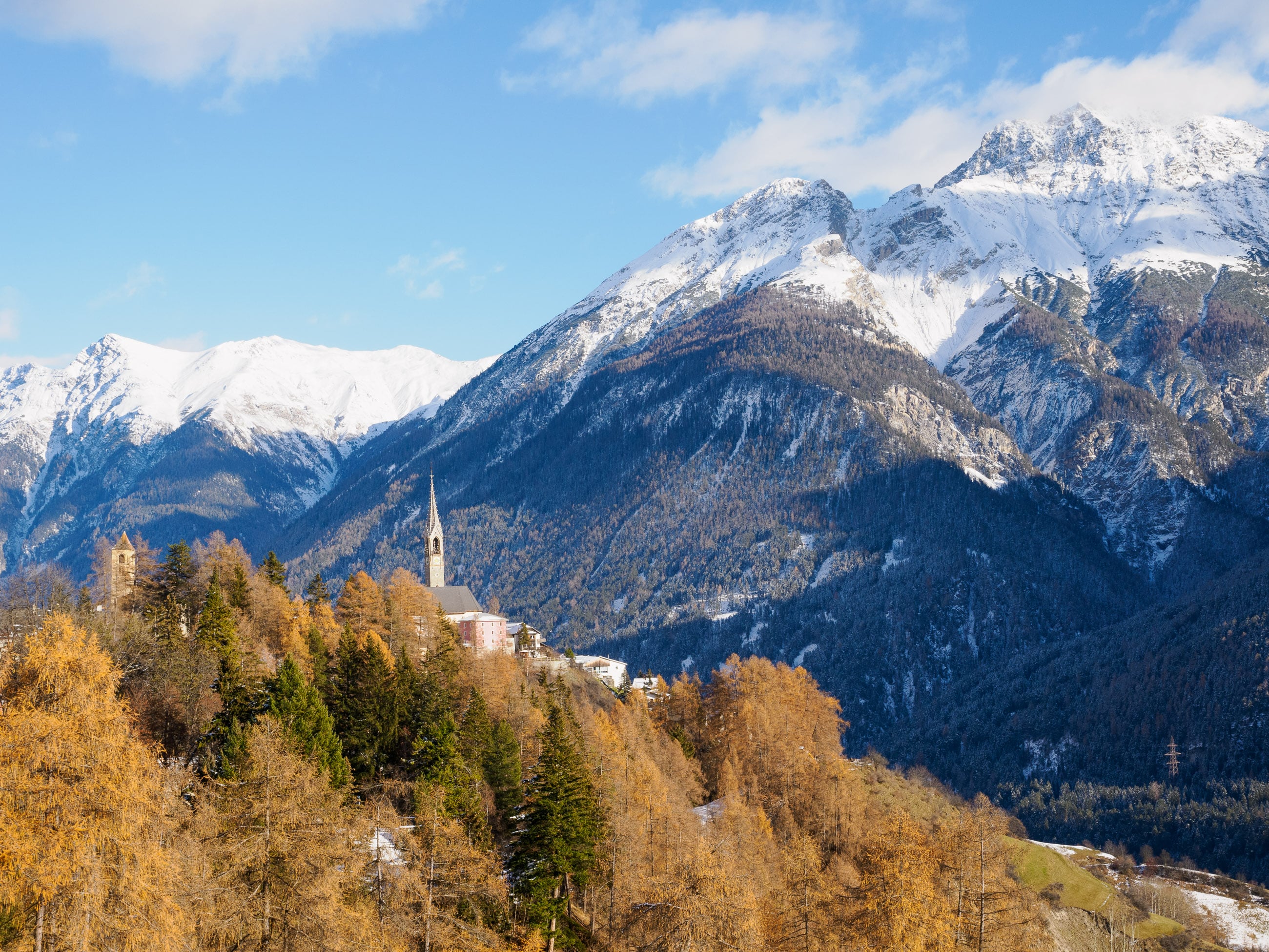 Village with tall spire on treed hillside next to snow-capped mountains