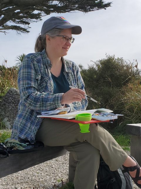 Melissa sitting on a bench with a sketchbook and watercolor palate on her lap while she looks into the distance with a brush poised above her paper