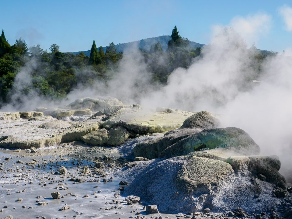 A landscape of gray rocks in the foreground, some stained yellow, with steam rising from it